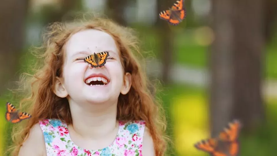 A girl is laughing. A butterfly sits on her nose.